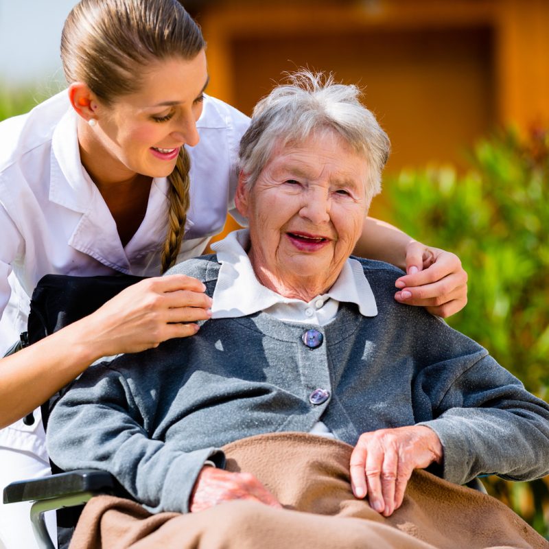 Senior woman in nursing home with nurse in garden sitting in wheelchair